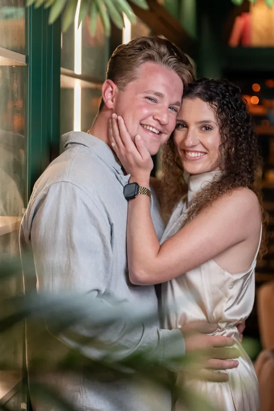 A person in a light blue shirt and another in a white dress embrace by a green pillar, their genuine joy evident.