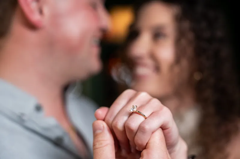 A close-up shows a hand displaying a diamond ring while the couple shares a joyful moment behind it.