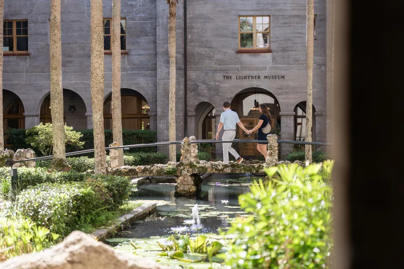 Two people stand together in the Lightner Museum Courtyard's lush garden, framed by tall palm trees and arched stone architecture.