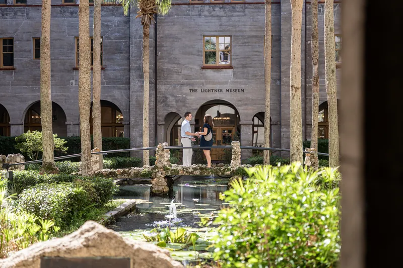 A person in a light blue striped shirt and another in a dark dress face each other across a wooden bridge in the museum's verdant courtyard.