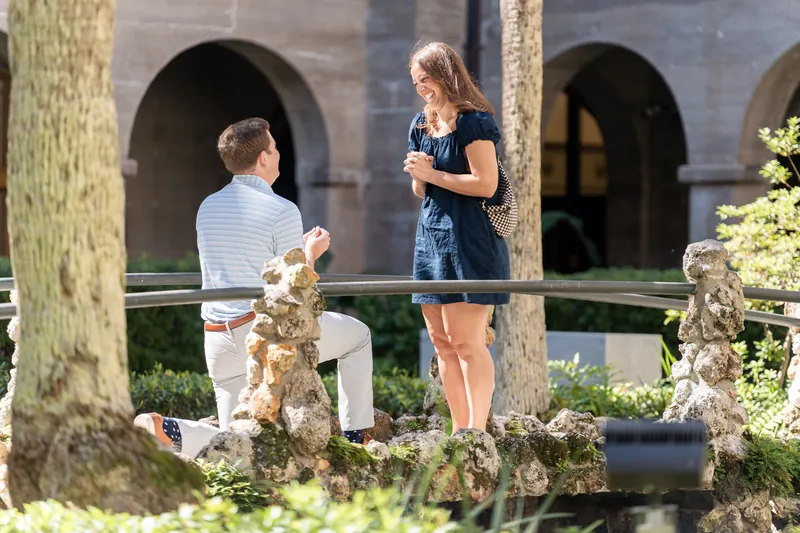 One person kneels on a wooden bridge while the other stands nearby, surrounded by lush vegetation and stone archways in the museum courtyard.