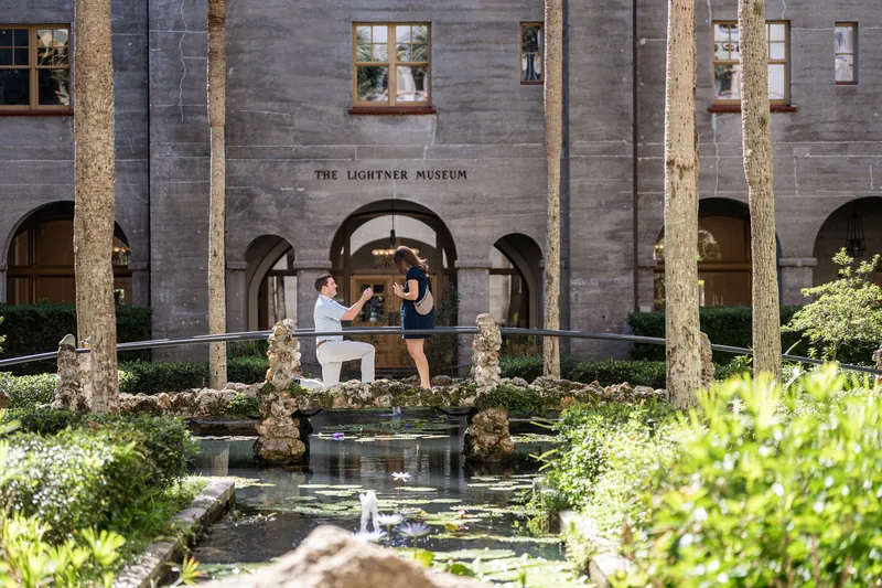 Two people embrace on a wooden footbridge beneath the stone arches of the Lightner Museum's courtyard.
