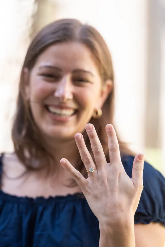 A woman in navy beams with her left hand raised, displaying a diamond engagement ring with the courtyard's arches softly blurred behind her.