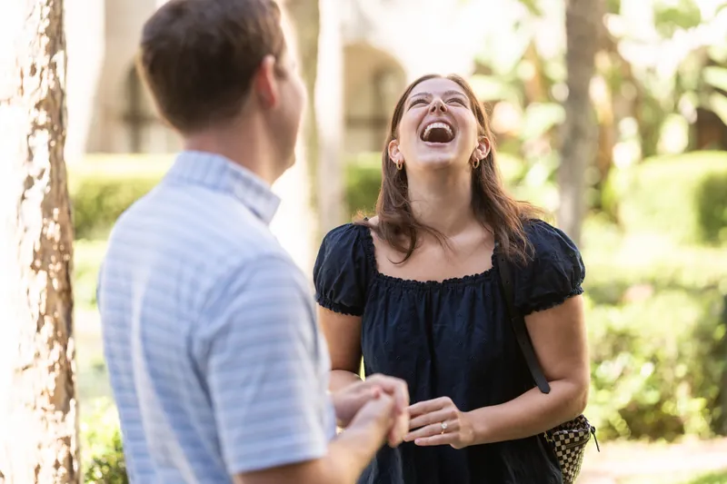 Two people laugh together beneath the museum's grand columns, their genuine joy evident as sunlight filters through the surrounding foliage.