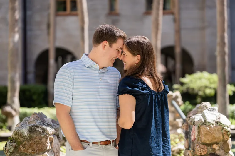 Two people lean in close, foreheads nearly touching, with stone planters and lush greenery visible in the courtyard setting.
