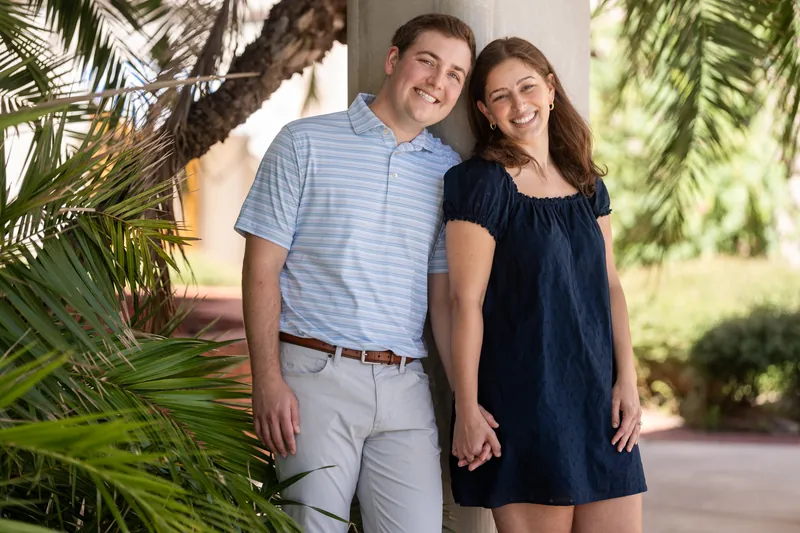 A man in a striped shirt and woman in a navy dress hold hands while walking through a shaded colonnade lined with tropical plants.