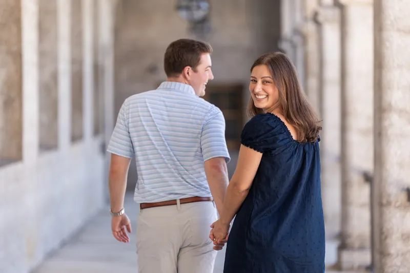 Walking along a stone corridor, the couple smiles at each other while holding hands, with soft natural light filtering across the archway.
