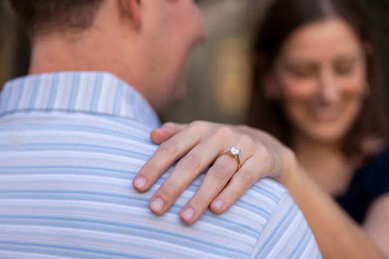 Close-up of a hand with a diamond ring resting on someone's shoulder in a light blue striped shirt.