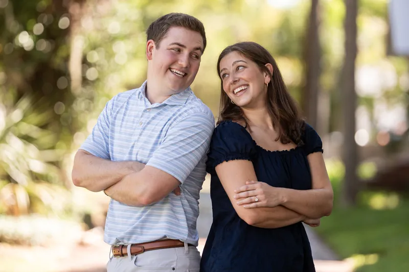 Both partners stand with arms crossed, laughing together on a sunlit residential street.