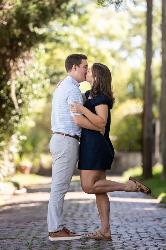 The couple shares a kiss on a quiet downtown street lined with green trees and historic architecture.