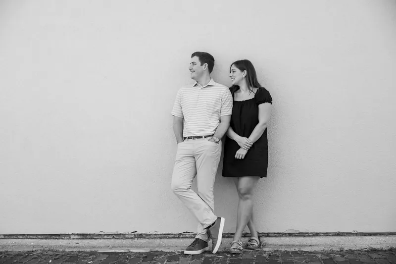 Two figures lean against a white wall in black and white, standing close and gazing toward each other.