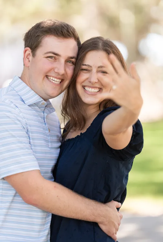 A smiling couple embraces outdoors with soft-focus greenery behind them, the person on the left wearing a striped shirt and the other in navy.