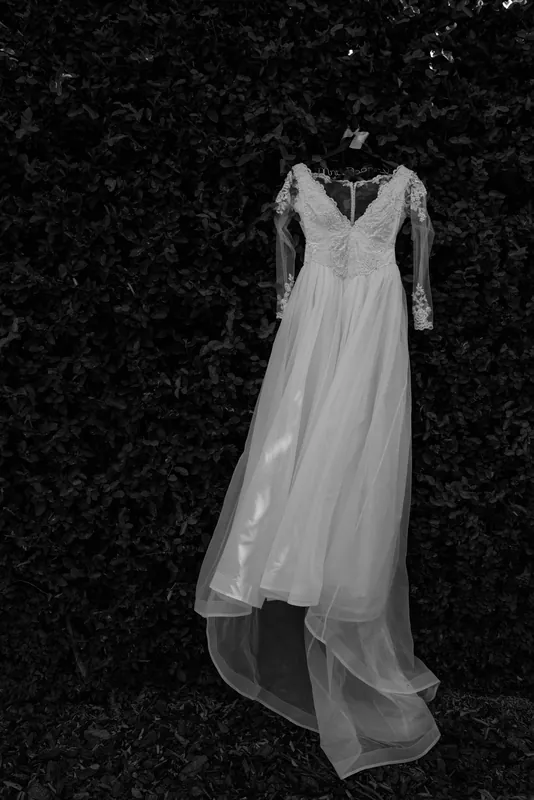Choko's white lace wedding dress with long sleeves and flowing train hangs against dark foliage in black and white.