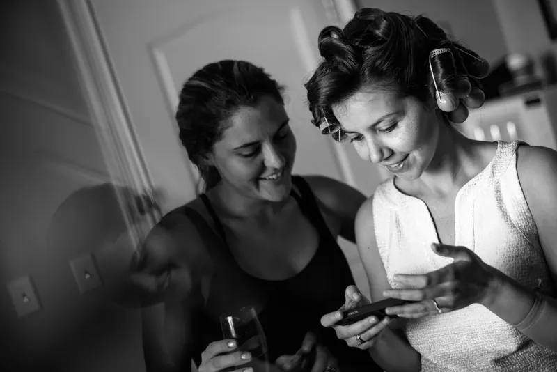 Two women look down together at Choko's phone, laughing during bridal preparations in black and white.