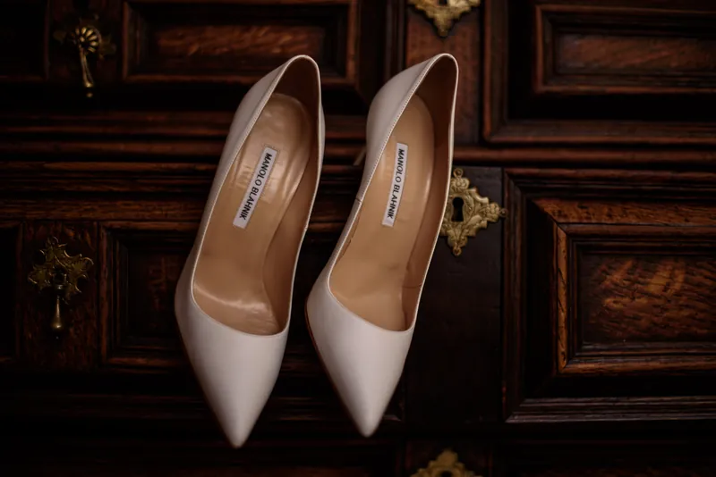 A pair of white pointed-toe heels sit on an ornate wooden surface with decorative gold accents.