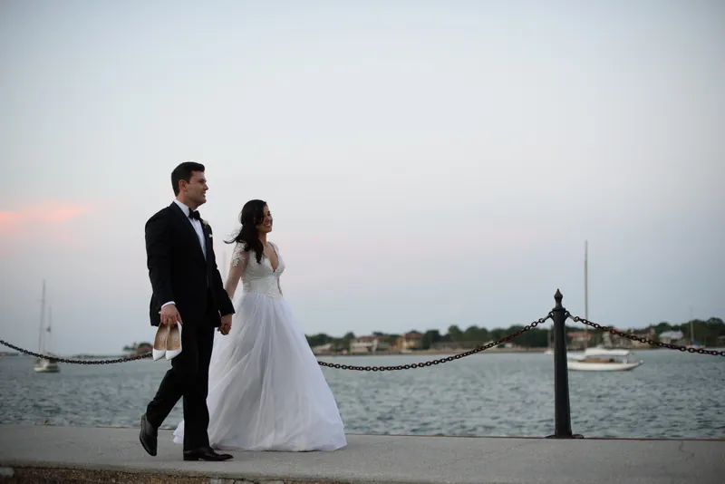 Choko walks arm-in-arm with Michael along a bayside promenade, both in wedding attire with the water and distant shoreline