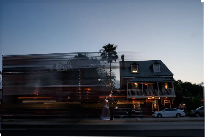A brick building with lit windows sits at dusk as a vehicle passes by on the street in front.