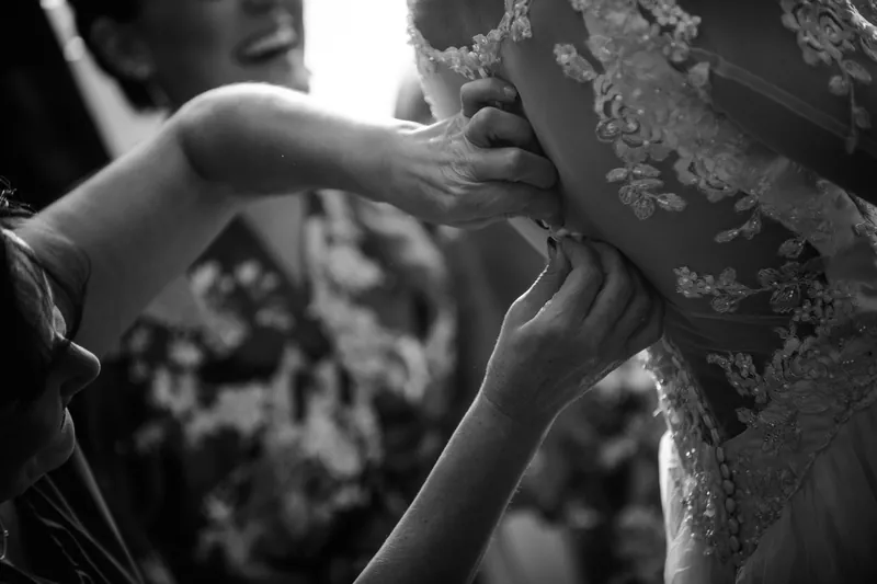 Someone adjusts the intricate beading on Choko's shoulder as she stands in her lace wedding dress.