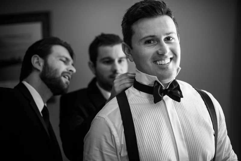 Michael grins at the camera wearing a bow tie and suspenders, flanked by two other men in formal wear.
