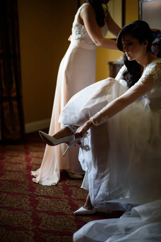 Choko lifts her flowing white gown as a woman helps arrange her train during bridal preparations.