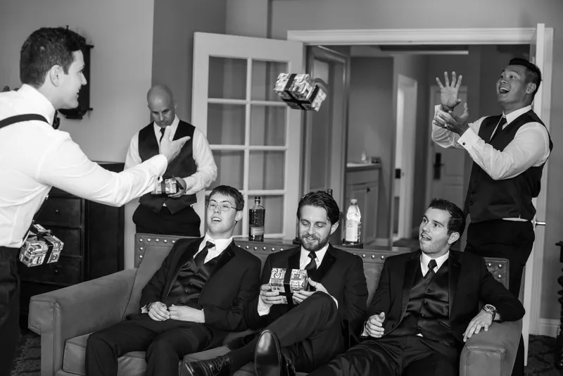 Groomsmen in vests and bowties laugh together while one man gestures animatedly during a candid moment indoors.