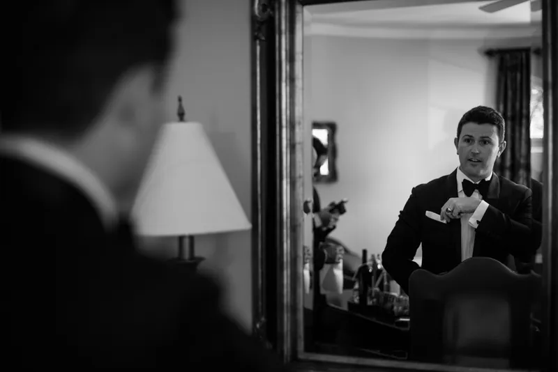Michael fastens his bow tie in a mirror, reflected against soft lamplight in the resort suite.