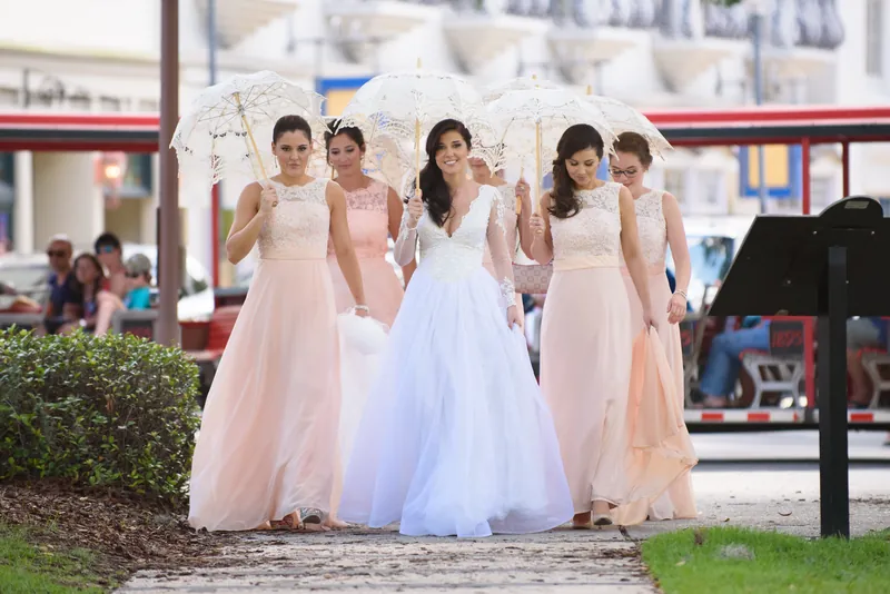 Choko in a white lace wedding dress holds hands with bridesmaids in blush gowns under a white lace parasol in Downtown St