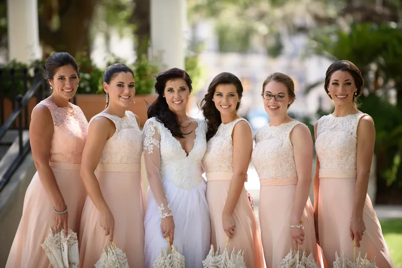 Six bridesmaids in matching blush and white gowns stand together outdoors in Downtown St Augustine, smiling at the camera.