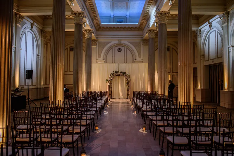 An elegant stone chapel with arched ceilings and glowing uplighting features rows of black chiavari chairs flanking