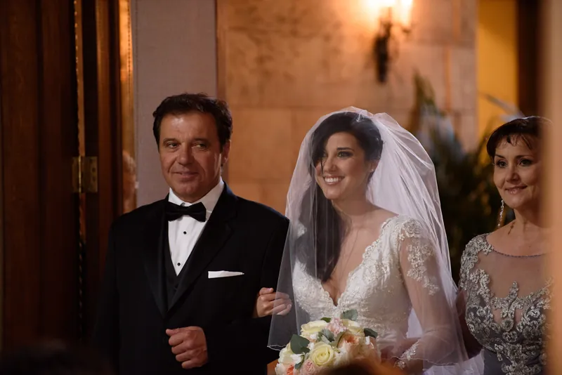 Michael in a black tuxedo escorts Choko down a warm-lit corridor, with a bridesmaid visible beside them.