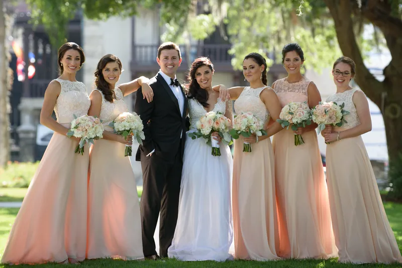 Seven bridesmaids in pink and blush dresses flank Michael and Choko on the grass, each holding a bouquet.