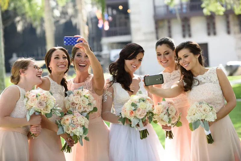Bridesmaids gather close, taking a selfie together while holding their pale pink and white flowers.
