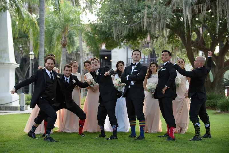 The wedding party erupts in playful poses across the sunlit lawn with Spanish moss-draped trees overhead.