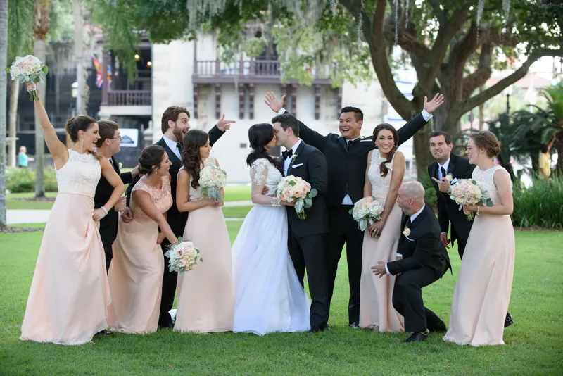 Choko and Michael stand arm-in-arm with their wedding party under oak trees, all smiling and holding bouquets.