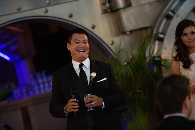Michael speaks into a microphone during toasts, wearing a black tuxedo with a boutonniere.