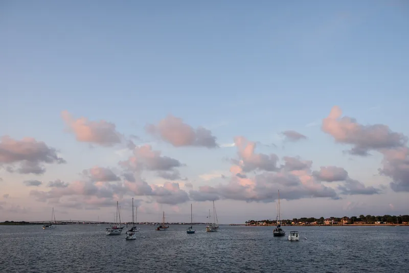 Sailboats dot the water at dusk under a pale sky with soft pink clouds above the St. Augustine waterfront.