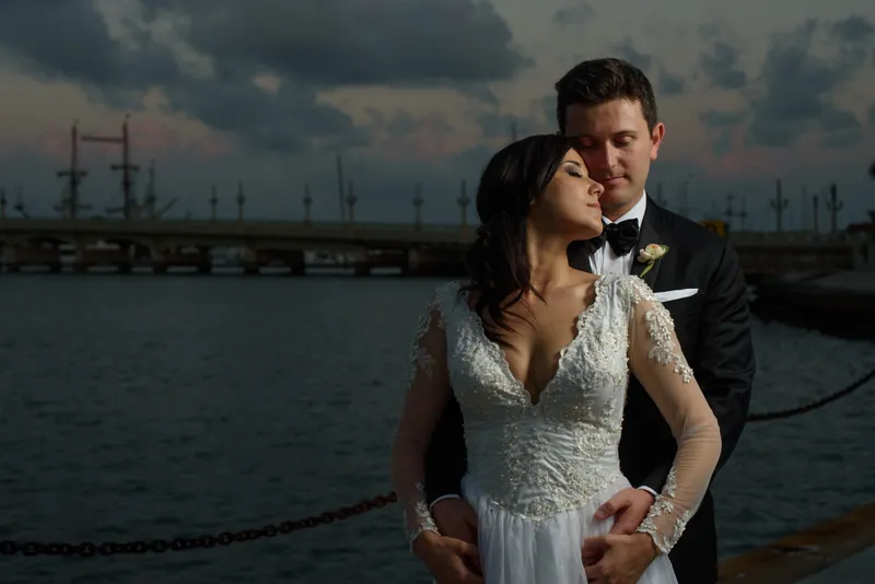 Choko and Michael stand close together by the water's edge, gazing toward the horizon with moored sailboats visible across