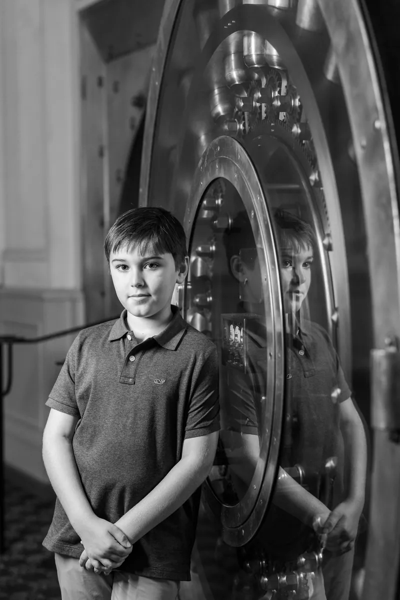 Black and white portrait of boy standing by Treasury vault door with reflection