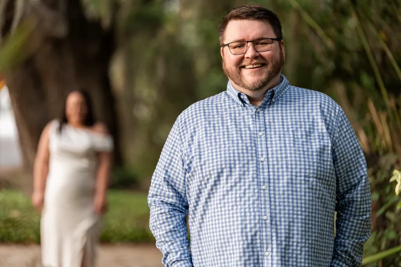 Chris smiles at the camera wearing a light blue checkered shirt while Shelby stands blurred in the background in a cream dress.