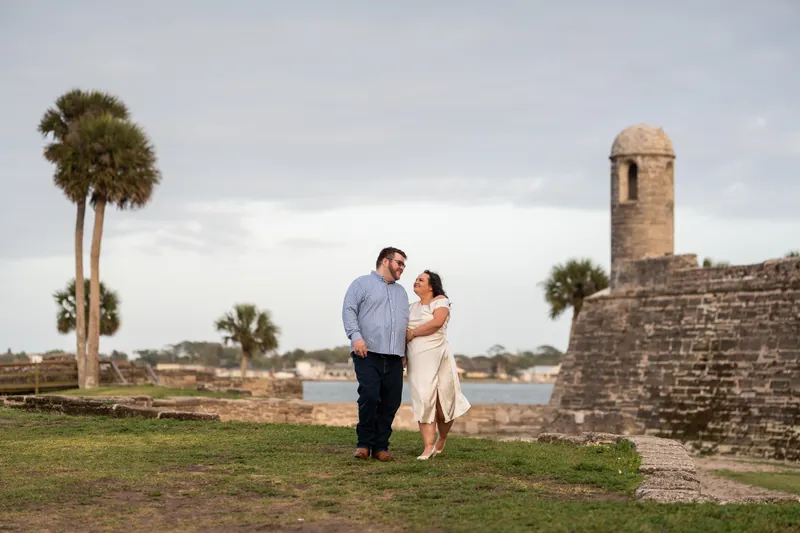 Chris and Shelby walk together on grass near a historic stone tower with a palm tree and cloudy sky behind them.