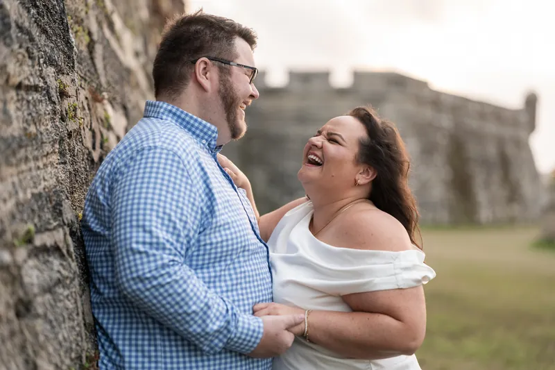 Chris and Shelby laugh together against a weathered stone wall with a fort visible in the background.