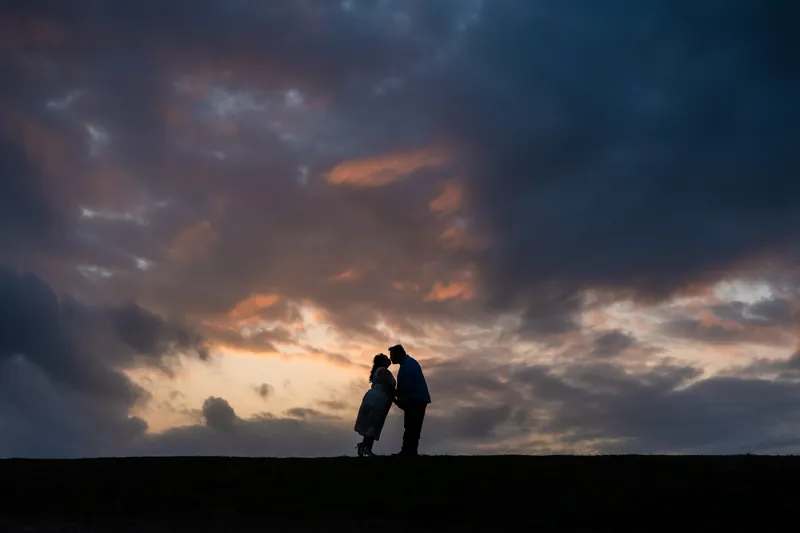 Chris and Shelby stand silhouetted against a dramatic sunset sky with orange and blue clouds.