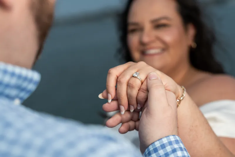 Shelby's hand rests on Chris's arm, displaying her engagement ring with water visible in the background.