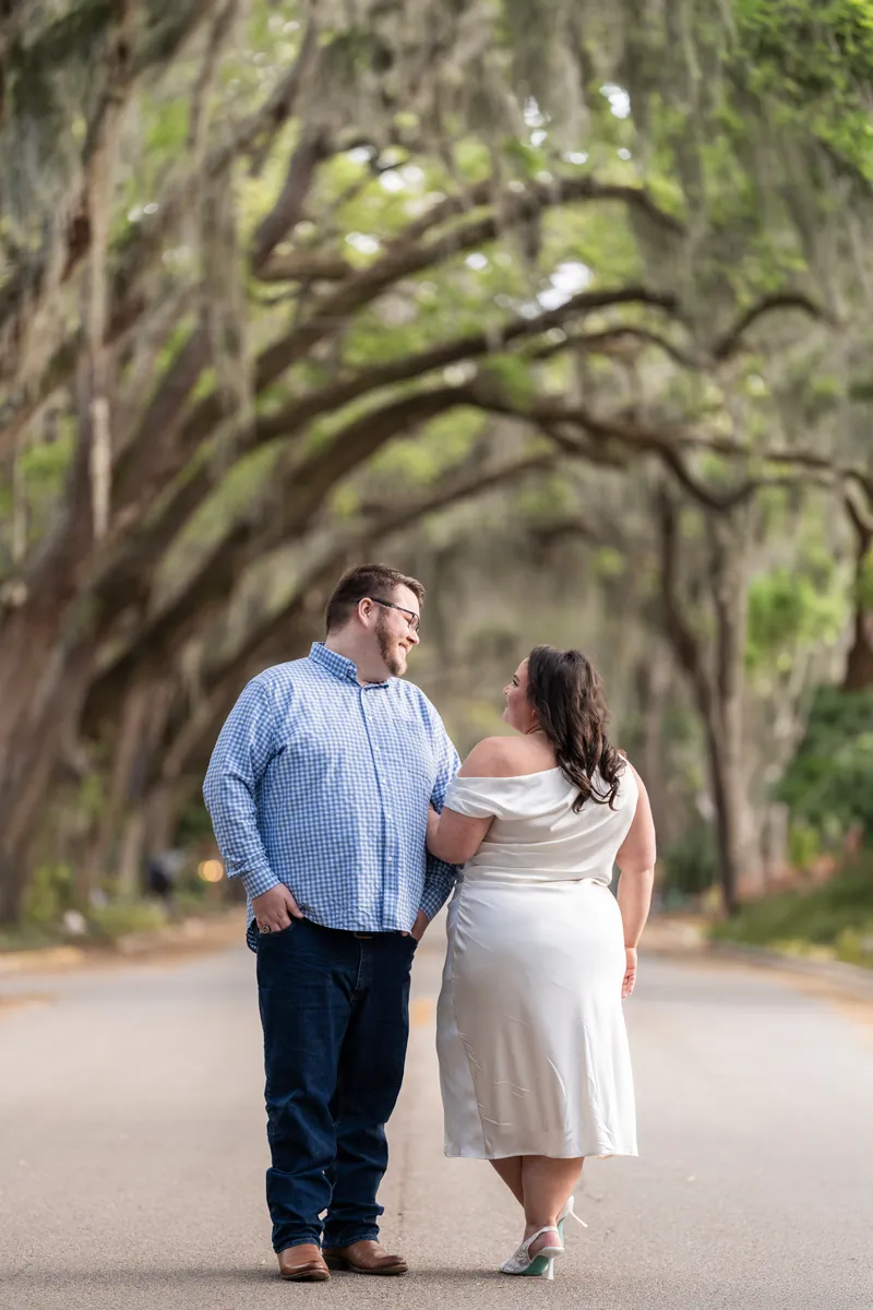 Chris and Shelby walk hand in hand down a tree-lined street beneath sprawling oak branches on Magnolia Avenue.