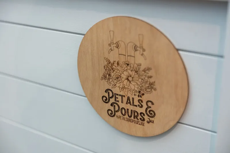 Wooden circular sign engraved with "Petals & Pours" and a floral design mounted on white shiplap.