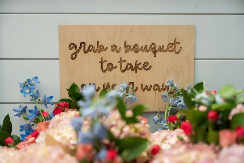 Wooden sign reading "Grab a bouquet to take your love away" surrounded by colorful flowers in soft focus.