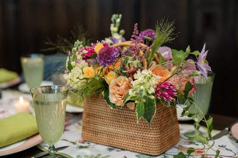 A coral and pink floral arrangement in a woven basket sits on a table setting with champagne glasses at the reception.