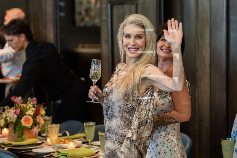 A blonde woman in a sparkly gray dress raises her champagne glass with a bright smile during the toast.