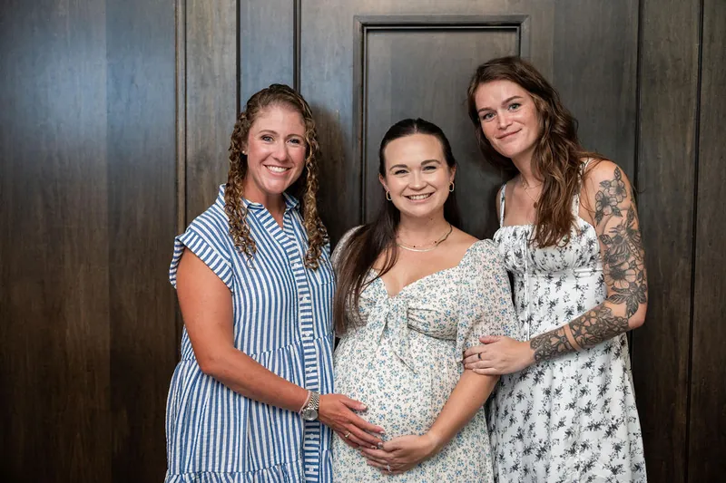 Three women stand together indoors, one cradling her pregnant belly while the others embrace her.