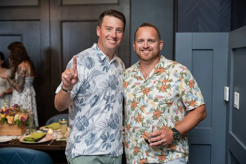 Two men in patterned short-sleeve shirts pose with arms around each other at the reception.